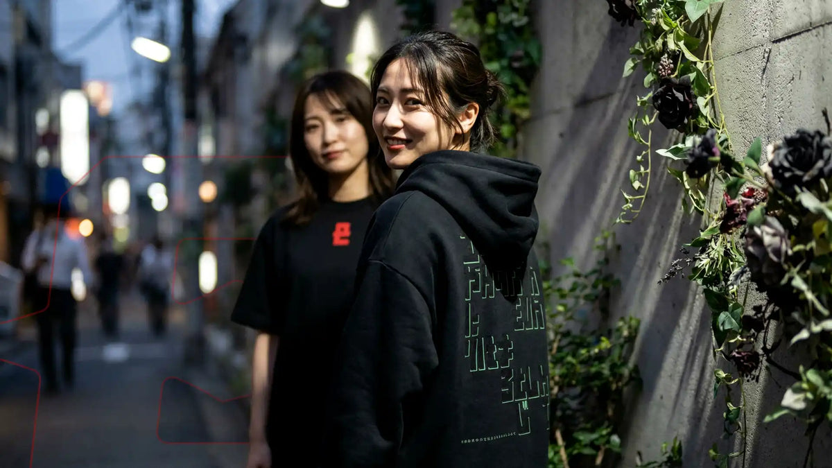 Deux femmes souriantes, l'une en t-shirt EXODE noir, l'autre en hoodie EXODE noir vue de dos, de nuit en ville