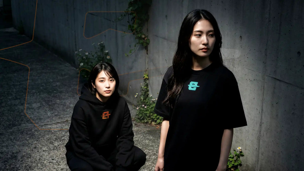 Deux femmes devant un mur en béton, l'une accroupie en hoodie EXODE noir, l'autre debout en t-shirt EXODE noir
